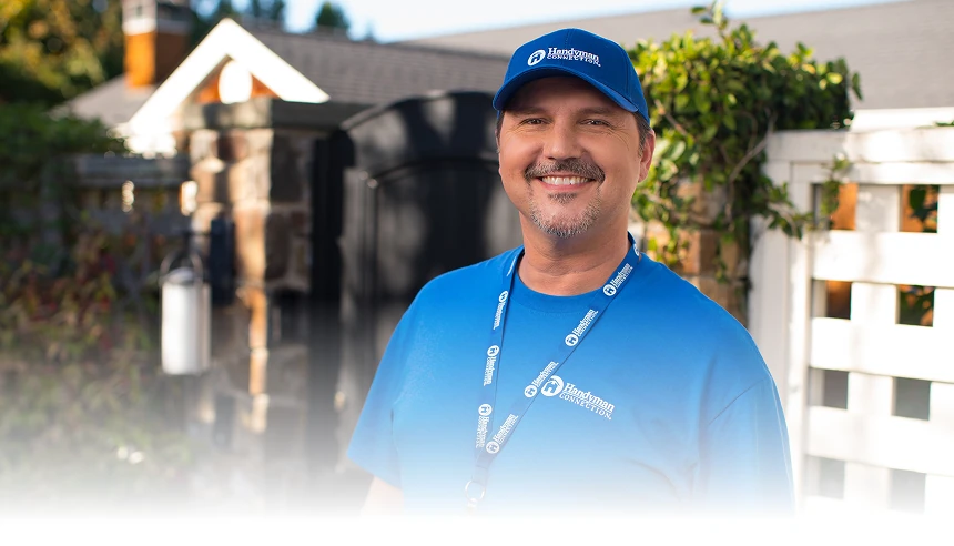 Smiling Handyman Connection employee in a blue company shirt, lanyard, and hat stands outside in front of a house, with the left side of the image fading to a blank white background.