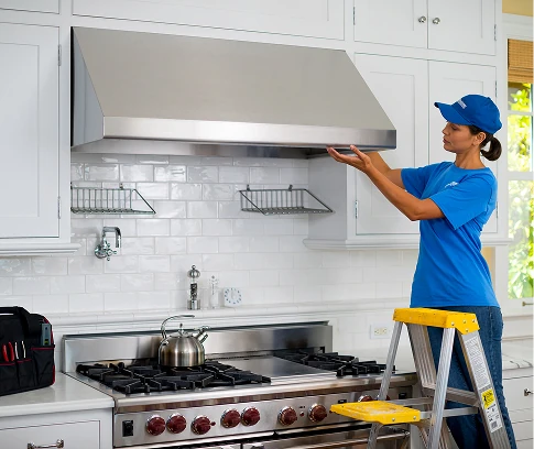 Technician in a Handyman Connection uniform stands on a step ladder and checks a large stainless range hood above a gas stove in a white kitchen.